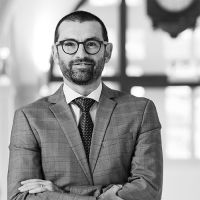 Thierry Omphalius, Directeur Administration et Finance, porte un costume sombre et une cravate à motifs, avec une expression légèrement souriante. Il se tient dans un cadre professionnel, avec un arrière-plan flou qui laisse entrevoir des éléments architecturaux modernes, créant une atmosphère sérieuse et professionnelle.