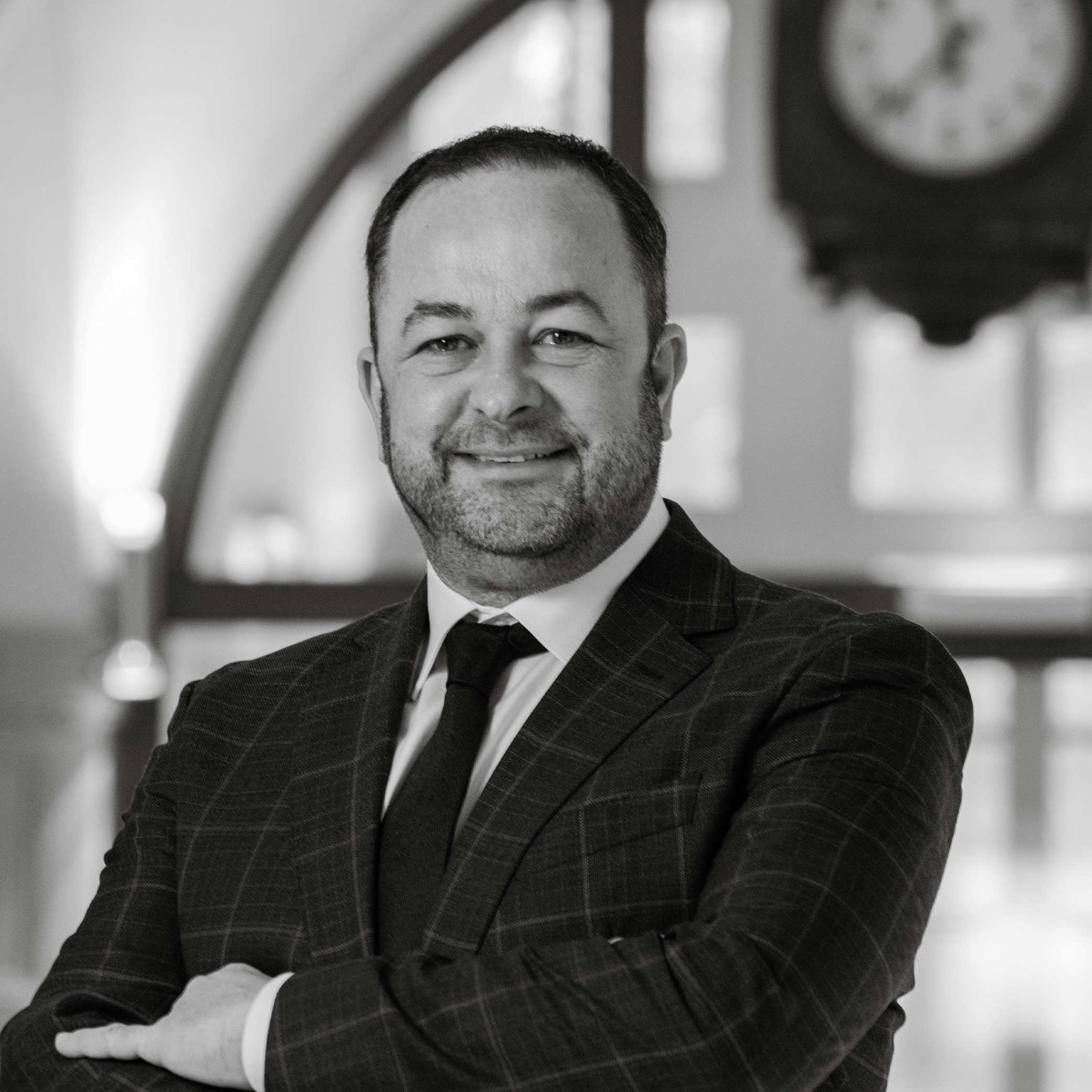 Carlo Diederich, Health & Spa Director, est représenté dans un portrait professionnel en noir et blanc. Il est debout, souriant légèrement, portant un costume foncé avec une cravate à rayures. Il porte des lunettes et son expression est calme et accueillante. L'arrière-plan est flou, montrant un hall avec une grande horloge en arrière-plan, ce qui ajoute une touche formelle et professionnelle à l'image.