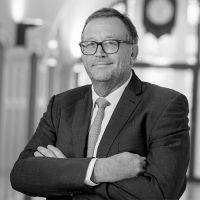 Pierre Plumer, Directeur Général, porte un costume sombre et une cravate rayée, avec une expression souriante. Il se tient dans un cadre professionnel, avec un arrière-plan flou qui montre une horloge murale, créant une ambiance sérieuse et accueillante.