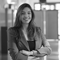 Anaïs Erpelding, Sales Manager, est représentée dans un portrait professionnel en noir et blanc. Elle est debout, souriante, les bras croisés, vêtue d'une veste élégante. L'arrière-plan est flou, montrant un environnement de bureau, ajoutant une touche professionnelle à l'image.