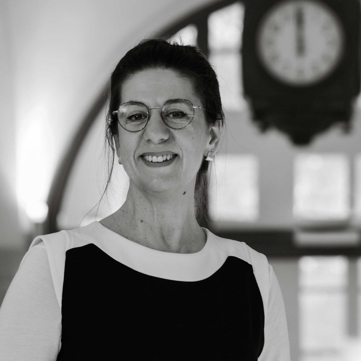 Kristina Guirguinova, est représentée dans un portrait professionnel en noir et blanc. Elle est debout, souriante, vêtue d'une robe noire et blanche. L'arrière-plan est flou, montrant un environnement de bureau, ajoutant une touche professionnelle à l'image.