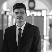Lucas Levrey, Events Manager, est représenté dans un portrait professionnel en noir et blanc. Il est debout, portant un costume sombre avec une cravate, et adopte une expression sérieuse mais accueillante. L'arrière-plan est flou, suggérant un environnement de bureau ou un hall d'entrée, ajoutant une touche professionnelle à l'image.