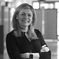 Marie-Anne François, Marketing & Communication Manager, est représentée dans un portrait professionnel en noir et blanc. Elle est debout, souriante, les bras croisés, vêtue d'un pull noir. Elle porte également une montre au poignet gauche. L'arrière-plan est flou, montrant un environnement de bureau, ajoutant une touche professionnelle à l'image.