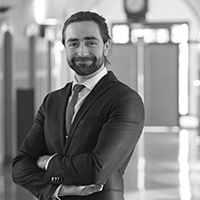 Yan Vullo, Hotel Manager, est représenté dans un portrait professionnel en noir et blanc. Il est debout, souriant, les bras croisés, vêtu d'un costume sombre avec une cravate claire. Il porte les cheveux attachés en arrière. L'arrière-plan est flou, montrant un environnement de bureau ou un hall, ce qui ajoute une touche formelle et professionnelle à l'image.