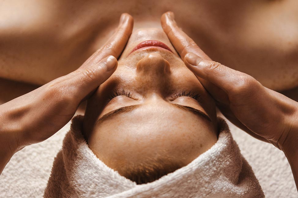 Frau liegt mit geschlossenen Augen und erhält eine Gesichtsmassage in einem Wellnessbereich.