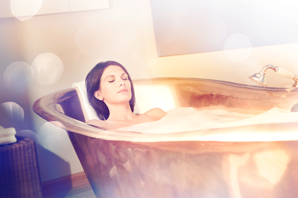 Une femme aux cheveux noirs est allongée dans une grande baignoire en cuivre, les yeux fermés, profitant d'un moment de détente. L'éclairage doux et les reflets dorés du cuivre créent une ambiance chaleureuse et apaisante, accentuée par des effets lumineu