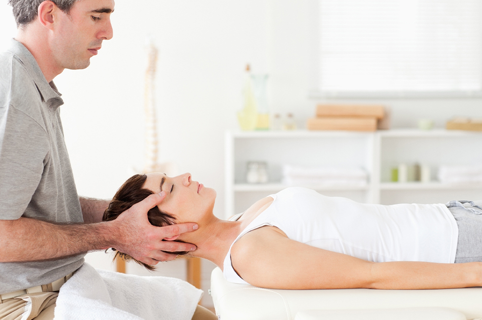 Un praticien effectue une thérapie craniosacrale sur une femme allongée sur une table de soin, soutenant doucement sa tête entre ses mains. L'environnement est lumineux et apaisant, avec des étagères en arrière-plan contenant des serviettes pliées et quel