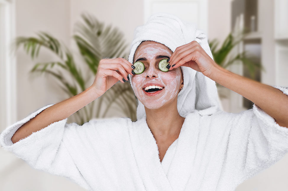 Femme en peignoir blanc avec un masque facial et des rondelles de concombre sur les yeux, souriante dans un environnement de spa lumineux.