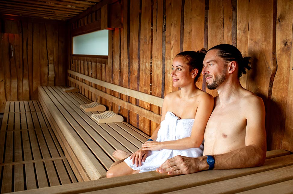 Couple assis dans un sauna en bois, profitant d’un moment de détente enveloppé dans une serviette blanche.