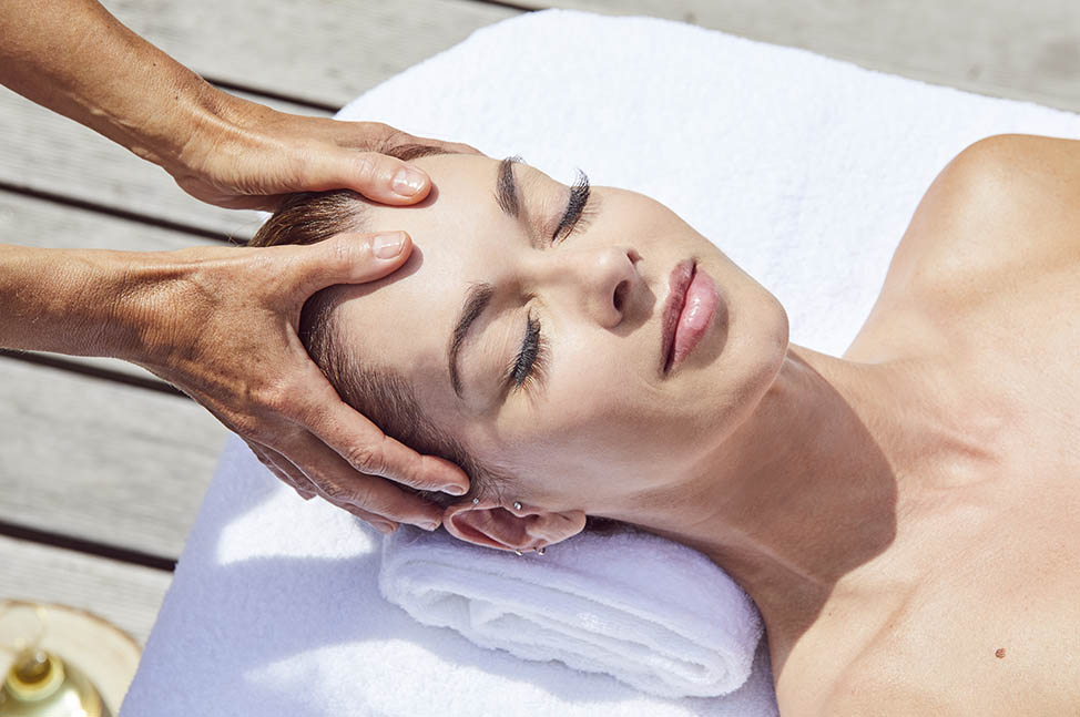 Une femme allongée, dans un cadre extérieur, reçoit un massage ayurvédique du visage et du cuir chevelu. Le praticien, à l’aide de mouvements doux et précis, masse délicatement la tête, procurant des effets apaisants et rajeunissants. Le cadre naturel ajo