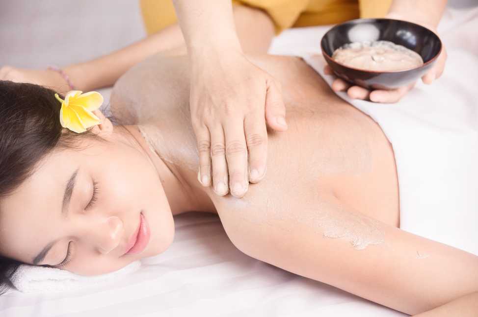 Une personne allongée sur le ventre reçoit un massage du dos dans un cadre de spa. Elle a une fleur jaune de frangipanier dans les cheveux et semble détendue. En arrière-plan, on aperçoit un bol contenant probablement des huiles ou des produits de massage