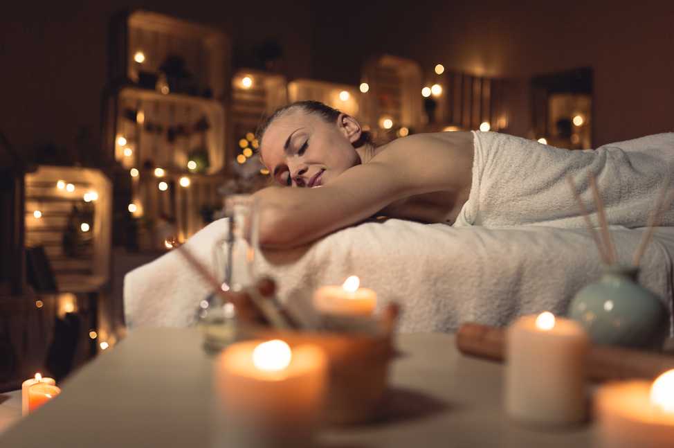 Une femme est allongée sur le ventre, couverte d'une serviette blanche, dans une pièce chaleureusement éclairée par des bougies. L'ambiance est apaisante et propice à la relaxation complète.