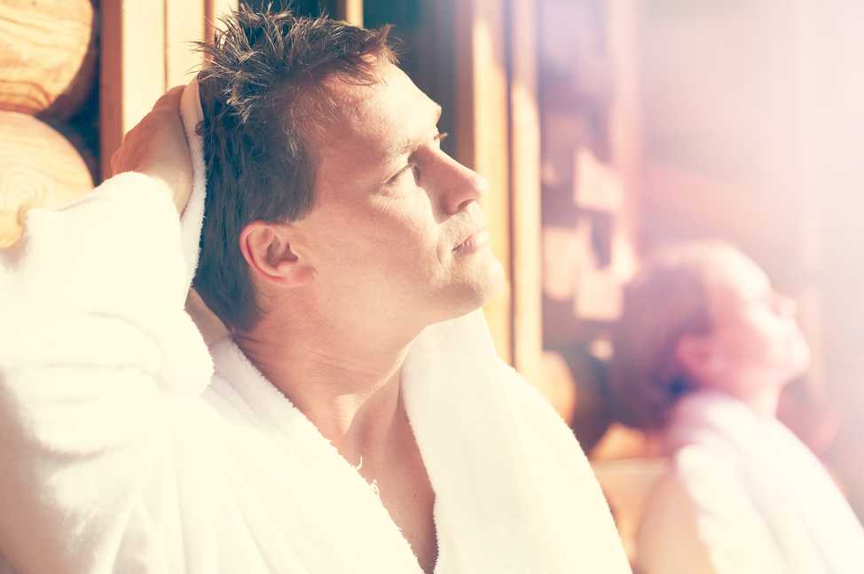 Un homme en peignoir blanc, utilisant une serviette sur sa tête pour s'essuyer ou se rafraîchir, profite d'un moment de relaxation dans un environnement lumineux et chaleureux en bois. En arrière-plan, une femme en peignoir est également en pleine détente